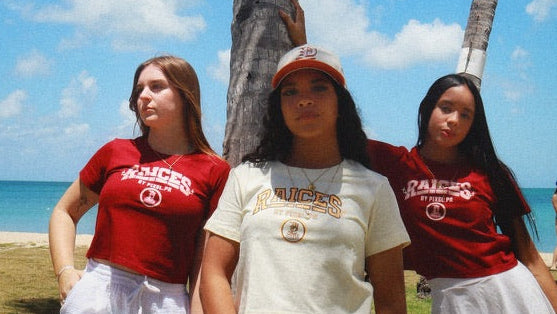 Three young women posing together outdoors under a palm tree on a grassy beach area with the ocean and blue sky in the background. Two of them are wearing matching red t-shirts with 'Cairns' printed on the front, paired with white shorts or skirts, and the center girl is wearing a cream t-shirt and white skirt.